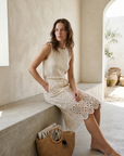 Femme assise, vêtue d'une jupe en dentelle crème en coton taille haute élégante et un haut confortable, dans un cadre minimaliste.