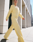 Modèle portant un ensemble deux pièces coton femme, composé d'une veste à col V en jaune et d'un pantalon assorti, marchant avec élégance dans un cadre moderne.