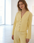 Femme souriante portant un ensemble deux pièces coton femme en jaune, avec une veste à col V et des boutons brillants, se tenant dans un intérieur lumineux.