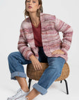 Femme portant un cardigan court en maille, coloris rose et beige, assise sur une chaise en osier. Ce cardigan court femme en maille a une coupe moderne avec des poches.