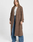 Manteau long femme en coton coupe ceinturée, porté par une femme devant un fond blanc, affichant un motif à carreaux marron avec pantalon large.