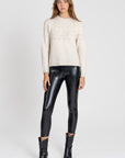 Femme portant un legging noir brillant taille haute, coupe ajustée, avec un pull beige en tricot et des bottes noires.