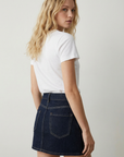 Femme montrant le dos de sa mini jupe en jean tendance, associée à un t-shirt blanc, dans un cadre simple.