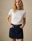 Femme souriante en mini jupe en jean tendance, posant devant un mur clair avec un t-shirt blanc.