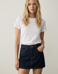 Modèle féminin en mini jupe en jean, style urbain, avec t-shirt blanc dans un décor minimaliste.