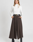 Femme portant un pantalon large taille haute pour femme, coupe fluide midi et ceinture intégrée, avec un haut blanc et des bottes noires.