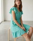 Robe fluide manches courtes en polyester légère — coupe féminine, modèle turquoise, assise sur une chaise moderne.