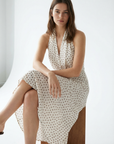 Femme portant une robe midi imprimée écru à pois, col halter décolleté plongeant, assise sur un tabouret en bois.