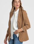 Femme portant un blazer femme chic crème, cascade de vêtements en arrière-plan pour un look branché.