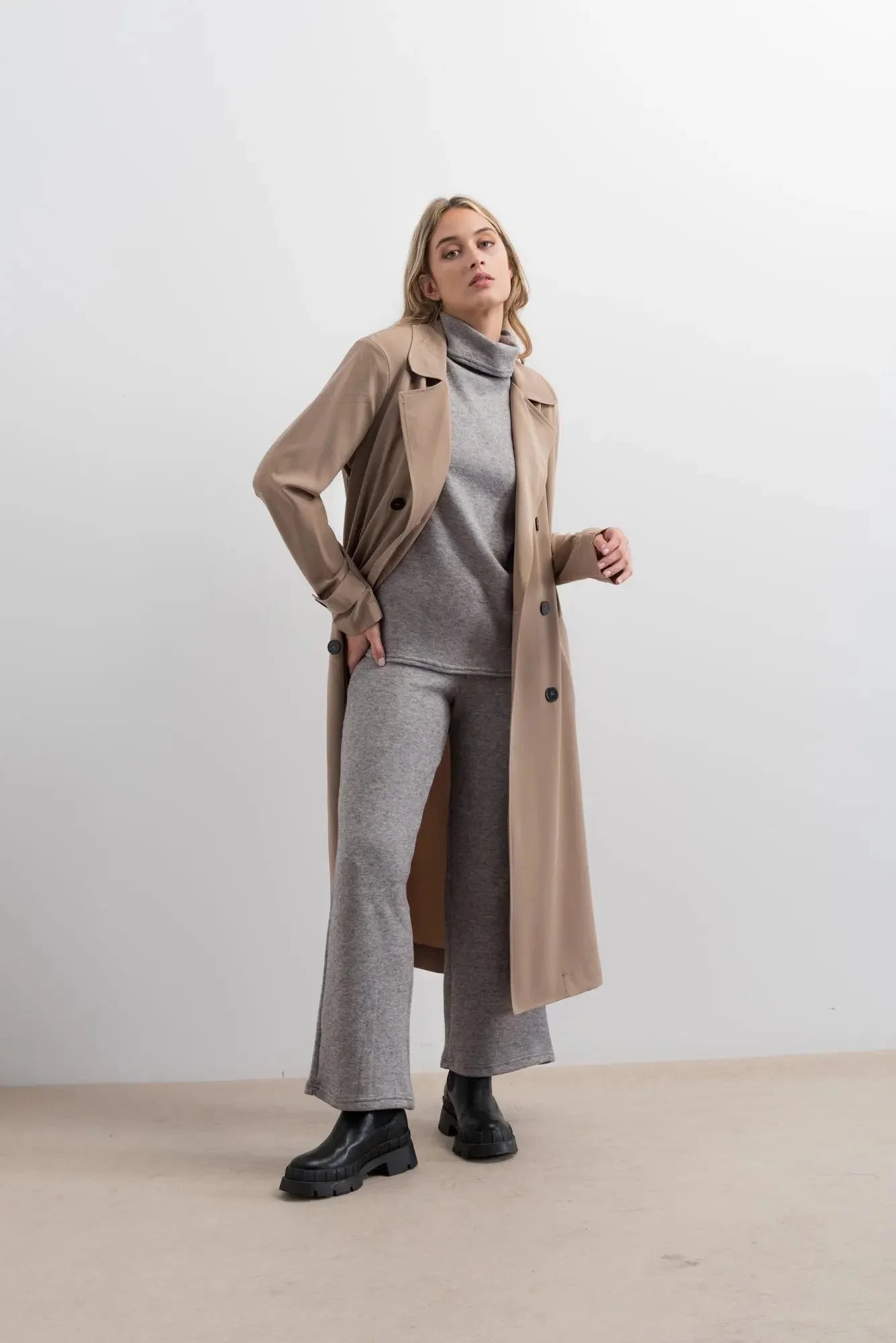 Vendeuse portant un trench beige long avec un col roulé gris et un pantalon large gris