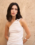 Haut femme blanc asymétrique avec boucle, porté par une femme aux cheveux bruns, fond beige. Élégance polyvalente pour chaque occasion.
