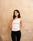 Haut femme blanc asymétrique porté par une jeune femme devant un mur beige avec un style décontracté. Élégance polyvalente pour chaque occasion.