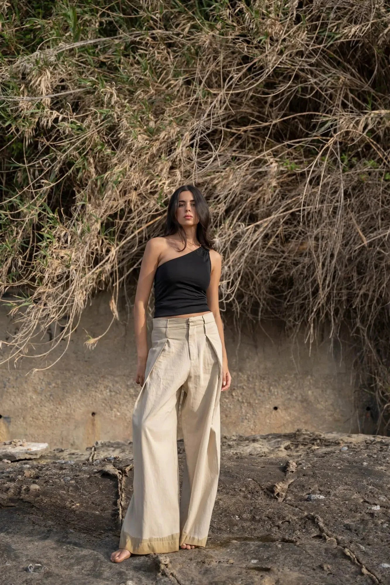 Femme posant près de la mer avec un haut asymétrique noir, portant un pantalon beige pour un look estival.
