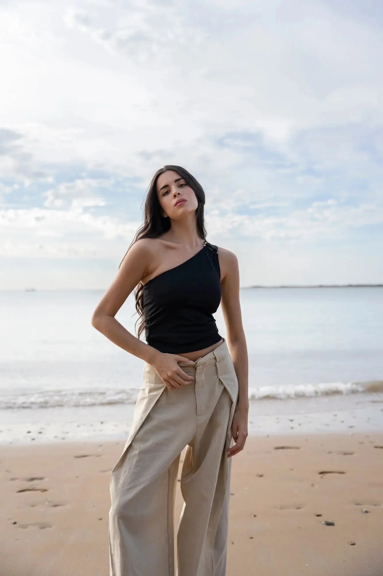 Femme debout sur la plage dans un haut asymétrique noir, ajoutant une sophistication à sa tenue estivale.