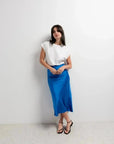 Femme portant la Jupe Satin Bleue Électrique Midi, élégante et flatteuse, associée à un haut blanc, idéale pour un look décontracté.