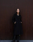 Un manteau long chaud femme noir à double boutonnage porté par une femme, avec un look chic et moderne, devant un mur en bois.