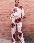 Femme souriante vêtue d'un ensemble décontracté dans un pantalon habillé fluide – Coupe droite moderne – Blanc à fleurs rouges – Matière légère, soulignant le pantalon motif femme.