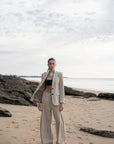 Femme élégante en pantalon large et fluide femme beige sur la plage, associée à une veste, style chic et décontracté.