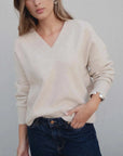 Femme portant un pull maille fine femme ample beige col V oversize chic et décontracté, avec un denim décontracté, devant un mur neutre.