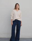 Mode femme avec un pull maille fine femme ample beige col V oversize, associé à un jean évasé, posant devant un fond beige.