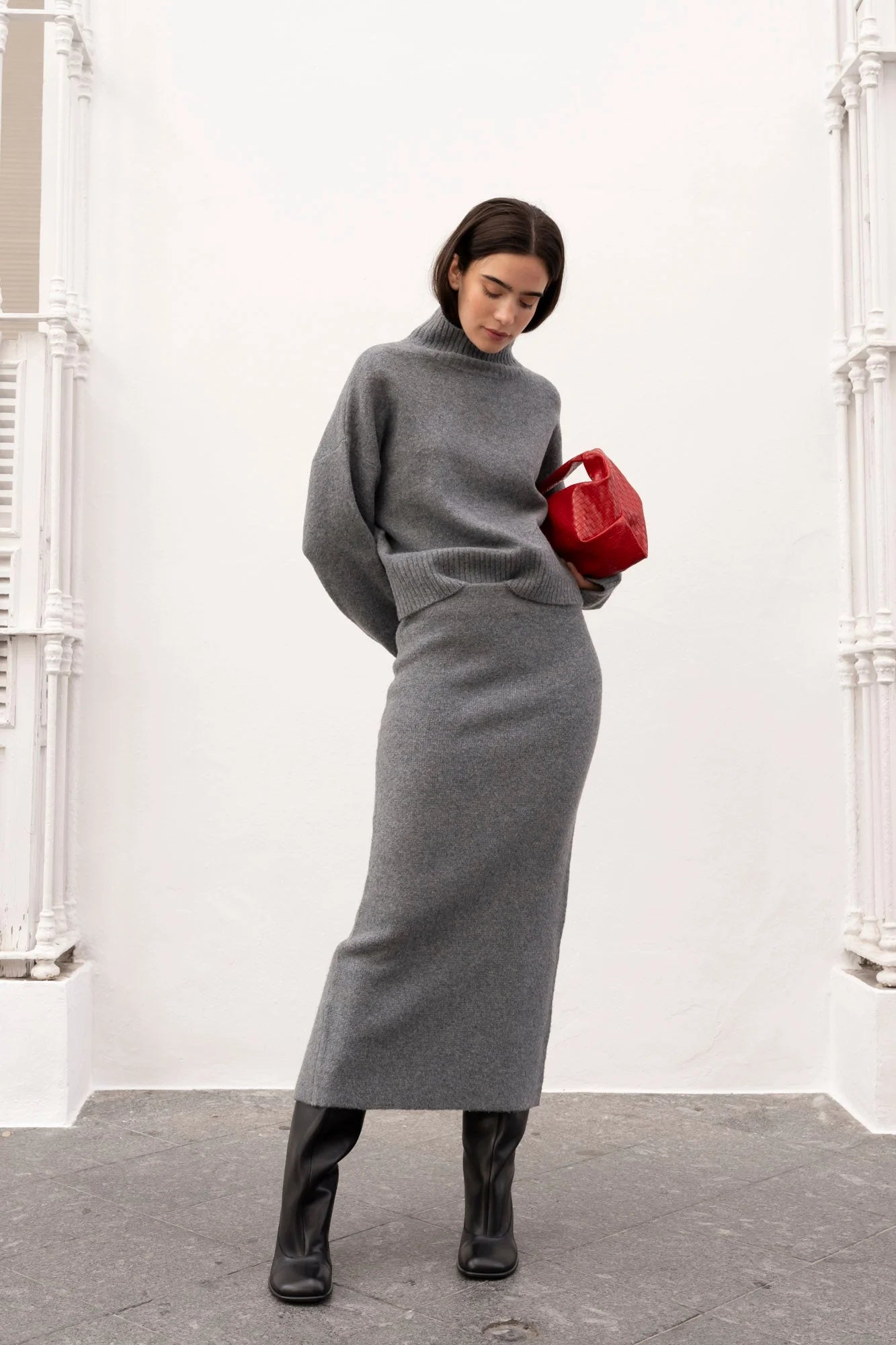 Pull court gris à col montant avec jupe midi, bottines noires et sac géométrique rouge
