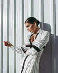 Femme posant dans une robe chemise longue chic à rayures noires et blanches, devant une surface métallique, montrant un look contemporain.