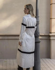 Femme vue de dos dans une robe chemise longue à rayures noires et blanches, s'appuyant contre un pilier, pour un look sophistiqué.