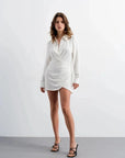 Modèle debout en Mini-Robe Portefeuille Chemise Courte Blanc Asymétrique - Élégance Chic et Structurée. La robe portefeuille blanche met en valeur la silhouette avec élégance.