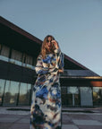 Femme posant avec assurance dans une robe satin longue manche longue aux motifs aquarelle, devant un design architectural contemporain, incarnant le style et le confort.