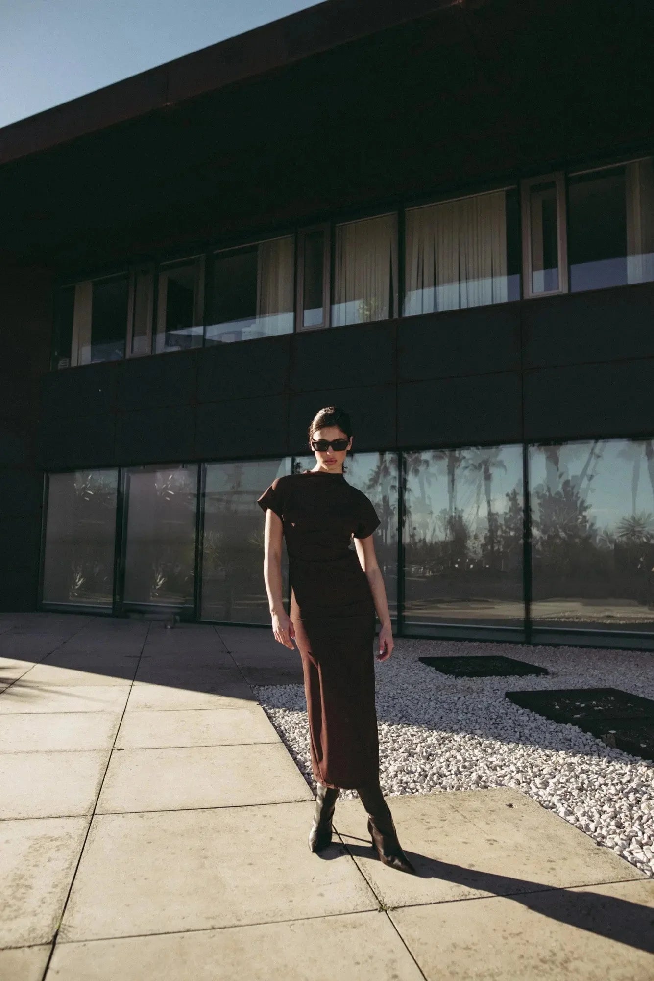 Robe longue marron ajustée femme avec détails drapés, modèle posant à l'extérieur avec des lunettes de soleil.