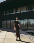 Robe longue marron ajustée femme avec détails drapés, modèle posant à l'extérieur avec des lunettes de soleil.