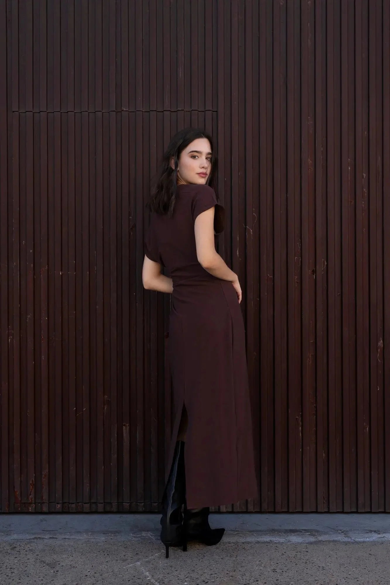 Robe longue marron ajustée femme avec détails drapés, vue de dos du modèle devant un mur en bois.