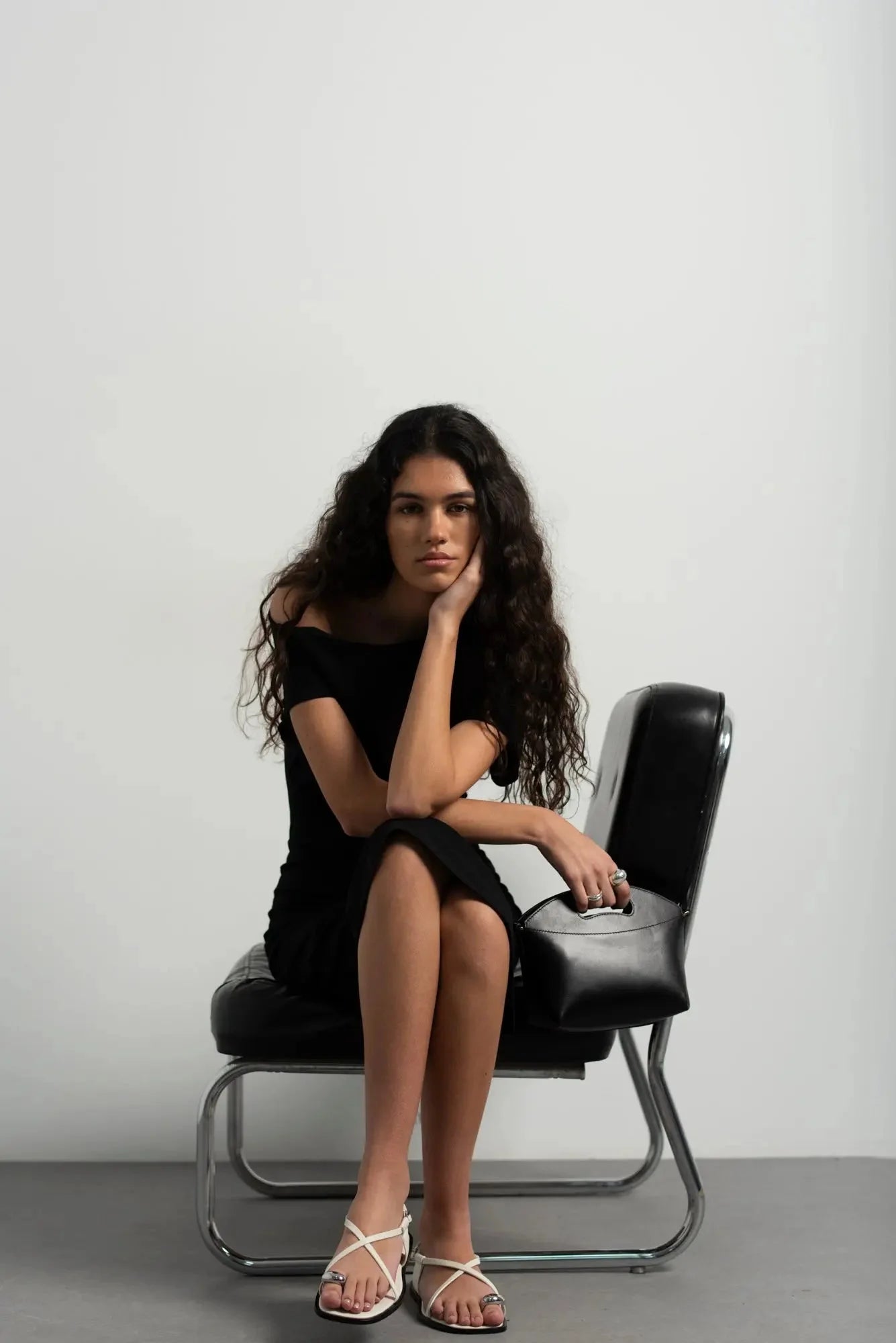 Femme en robe midi asymétrique pour soirée noire, assise avec sac noir et sandales blanches