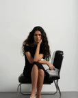 Robe asymétrique mi longue noire femme, élégance versatile pour soirées tendance 2024. Femme assise dans une pose réfléchie, mettant en valeur la robe élégante et raffinée.