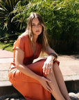 Femme assise dans un jardin, vêtue d'une robe midi satinée orange cuivré,款表现出te l'élégance naturelle de cette robe satin orange.