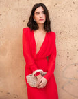 Femme portant une robe mousseline rouge midi à manches longues, décolleté plongeant élégant, accompagnée d'une pochette, parfaite pour des événements.