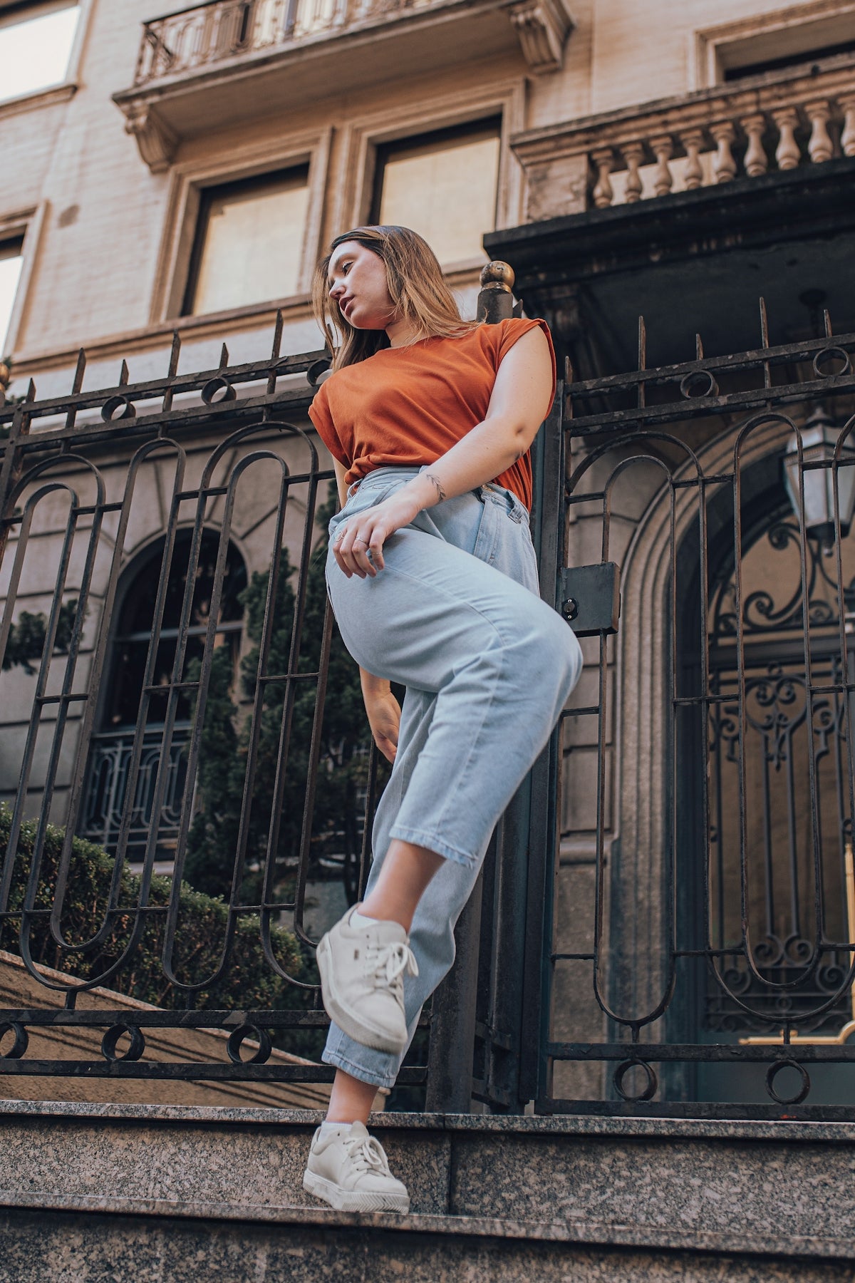 woman in orange t-shirt and blue denim jeans sitting on black metal fence during daytime