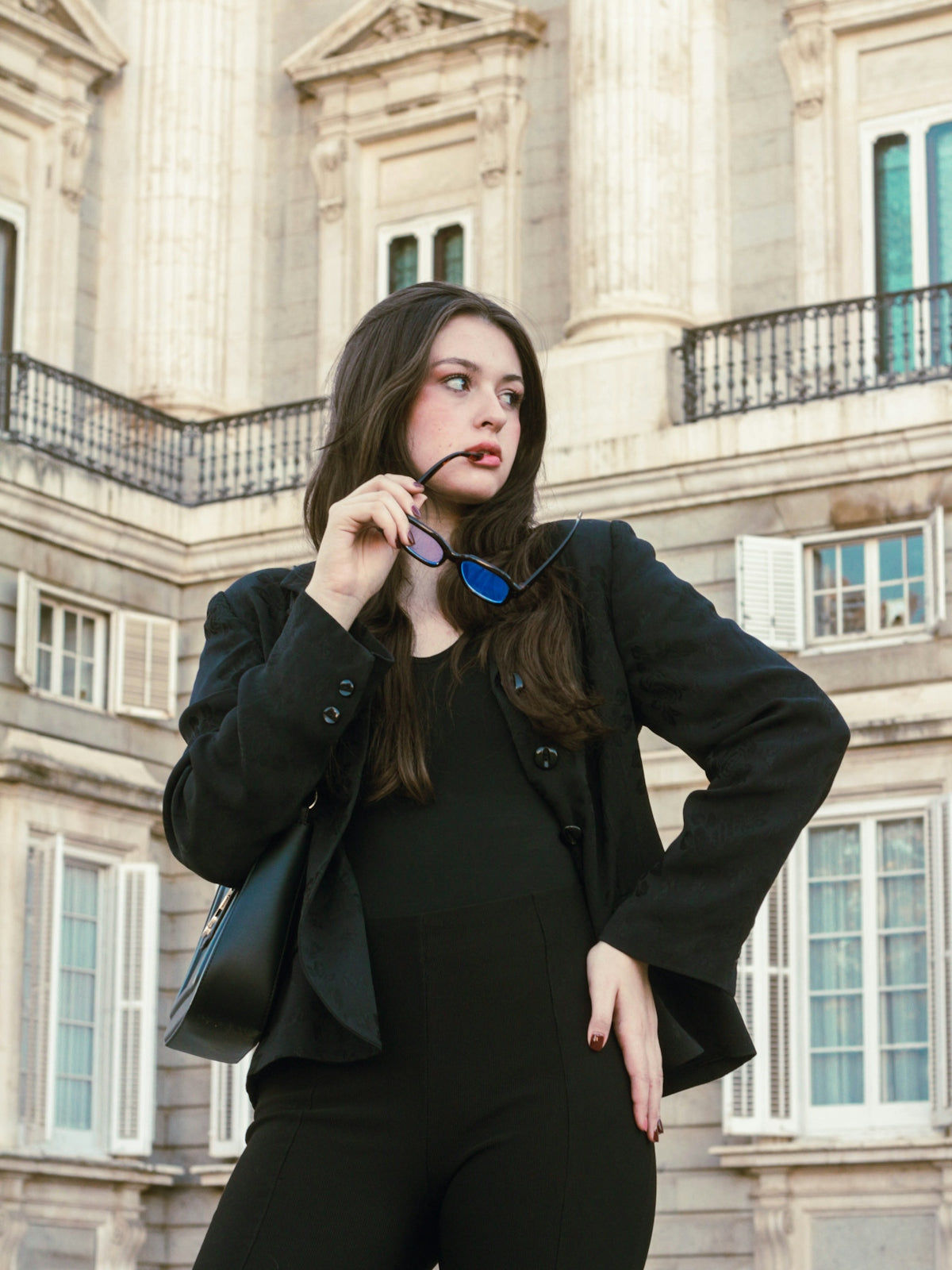 Woman in black outfit holds sunglasses near building