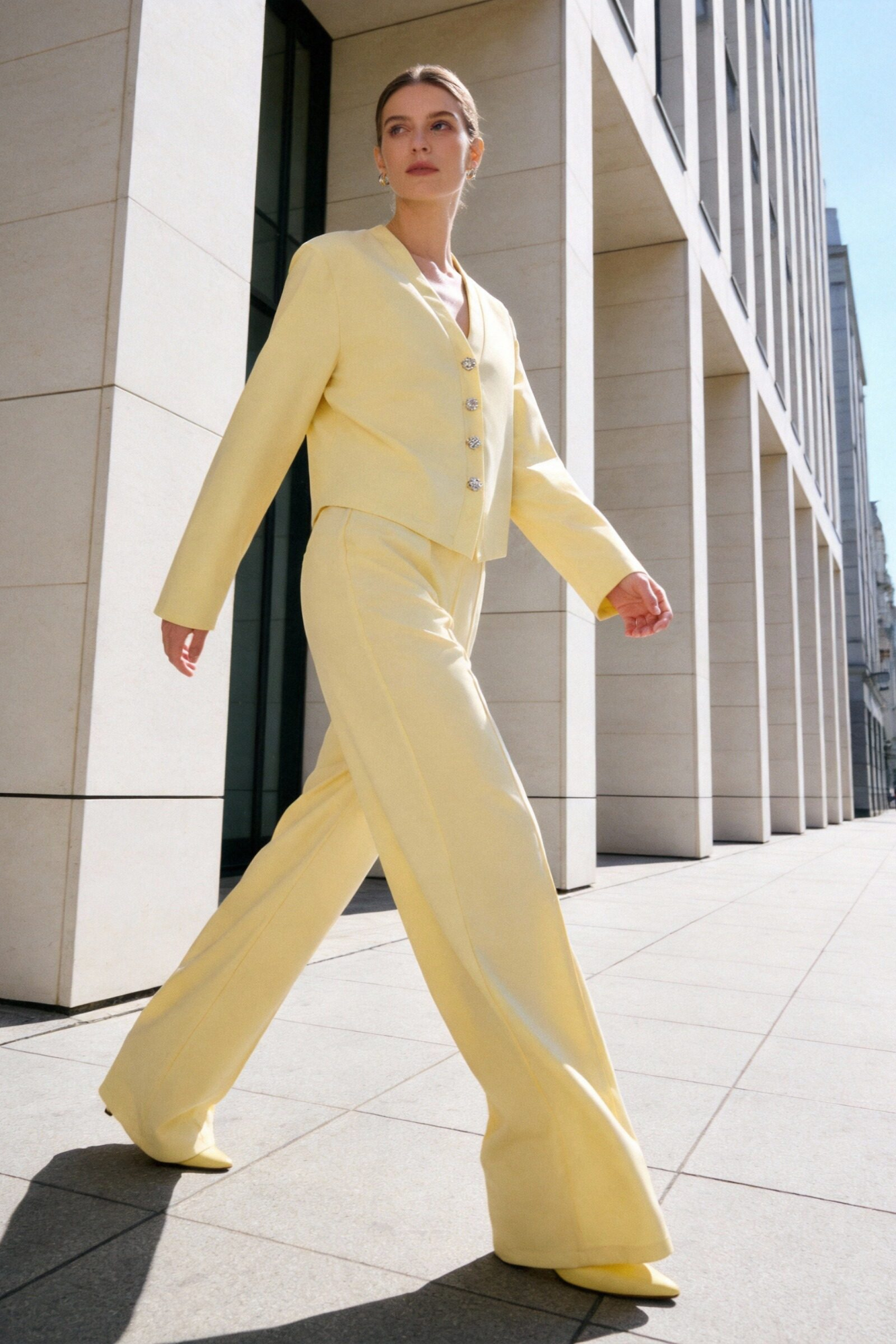 Modèle portant un ensemble deux pièces coton femme, composé d'une veste à col V en jaune et d'un pantalon assorti, marchant avec élégance dans un cadre moderne.