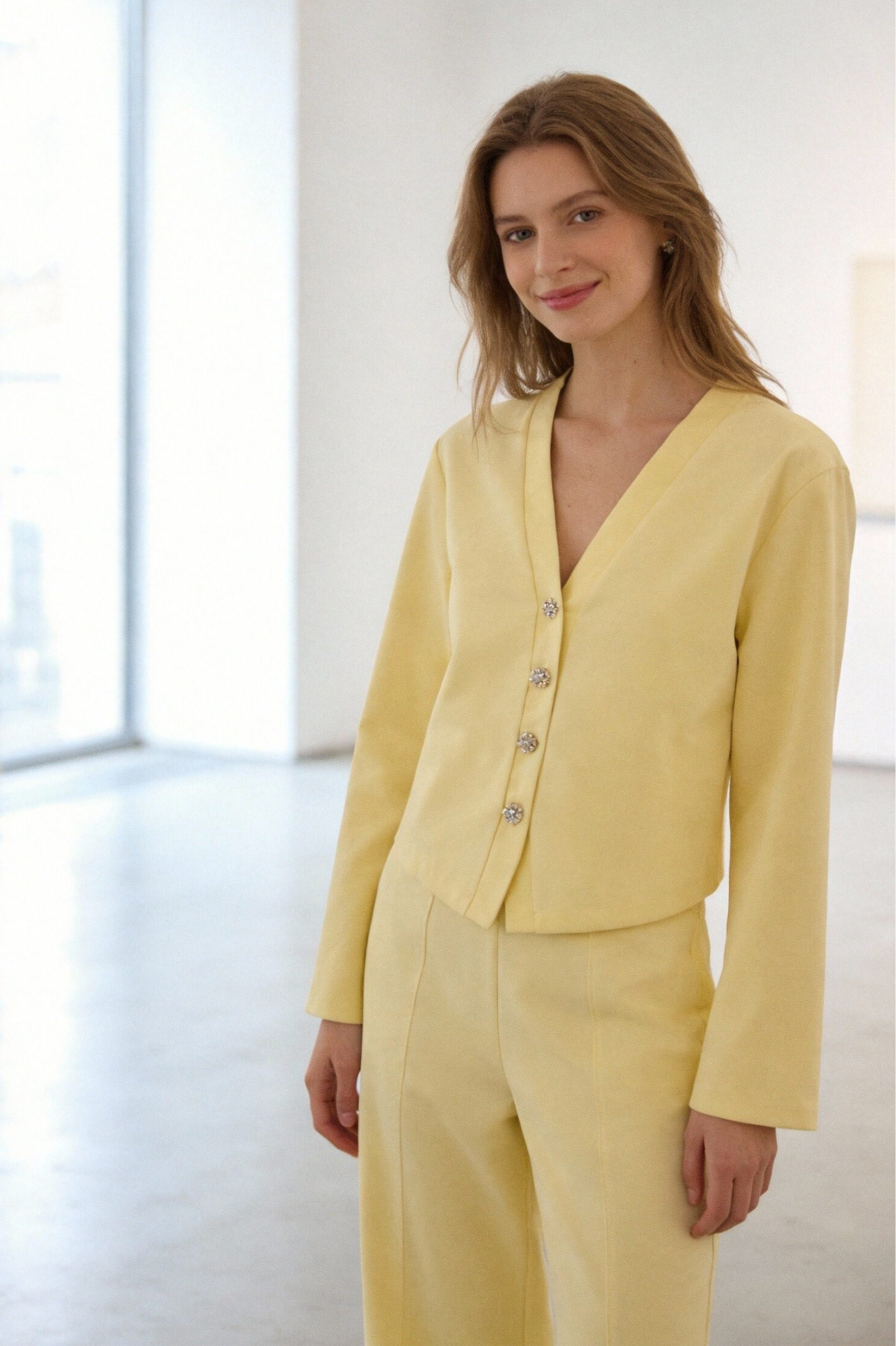 Femme souriante portant un ensemble deux pièces coton femme en jaune, avec une veste à col V et des boutons brillants, se tenant dans un intérieur lumineux.