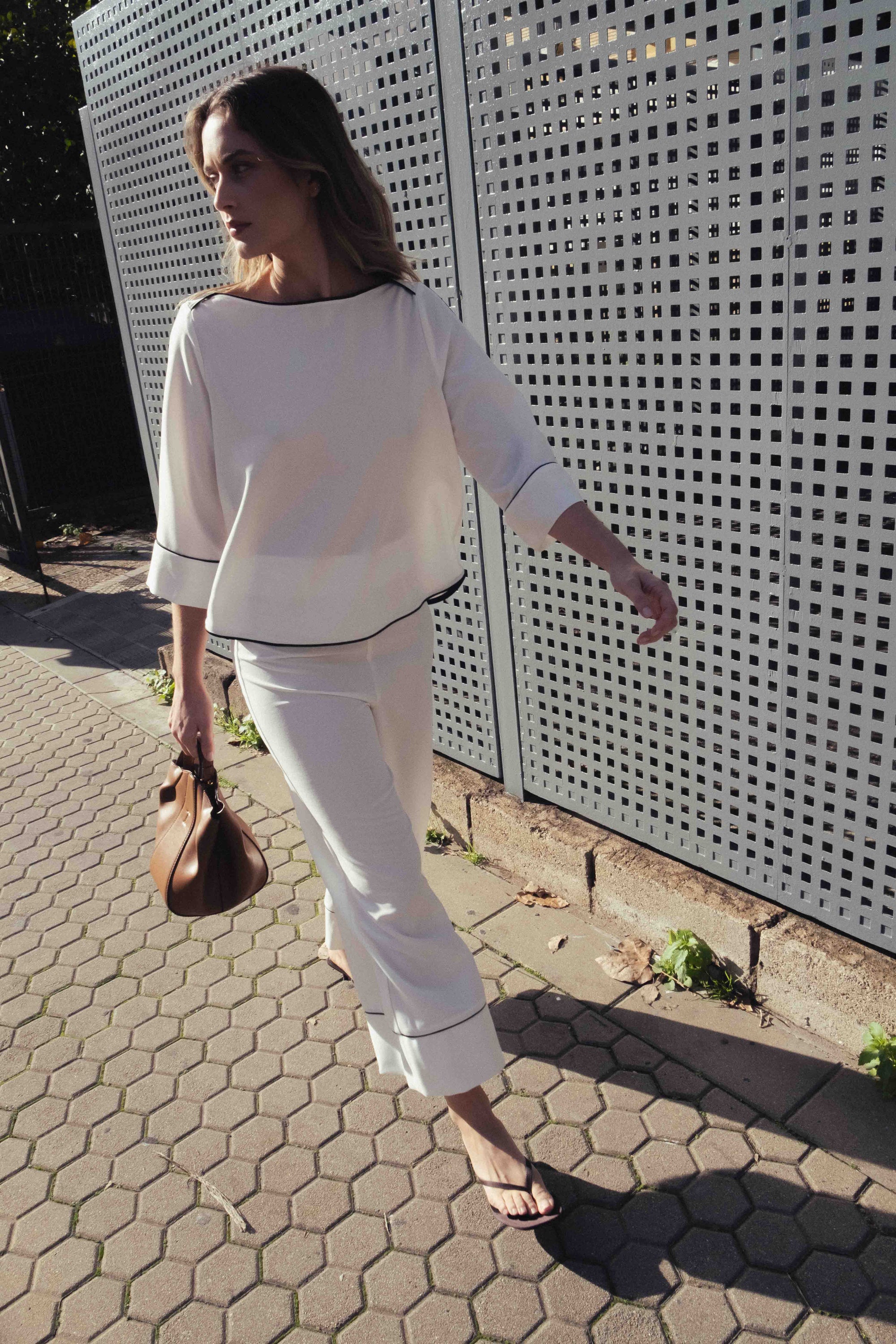 Femme portant un pantalon culotte écru taille mi-haute à bandes contrastées, marchant élégamment avec un sac à main, mettant en avant son style moderne et chic. Idéal pour un look de pantalon femme taille mi-haute.