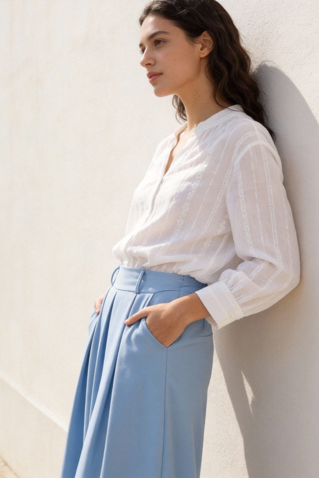 Femme avec un pantalon fluide taille haute femme bleu et une chemise blanche, posant contre un mur, look décontracté et élégant.