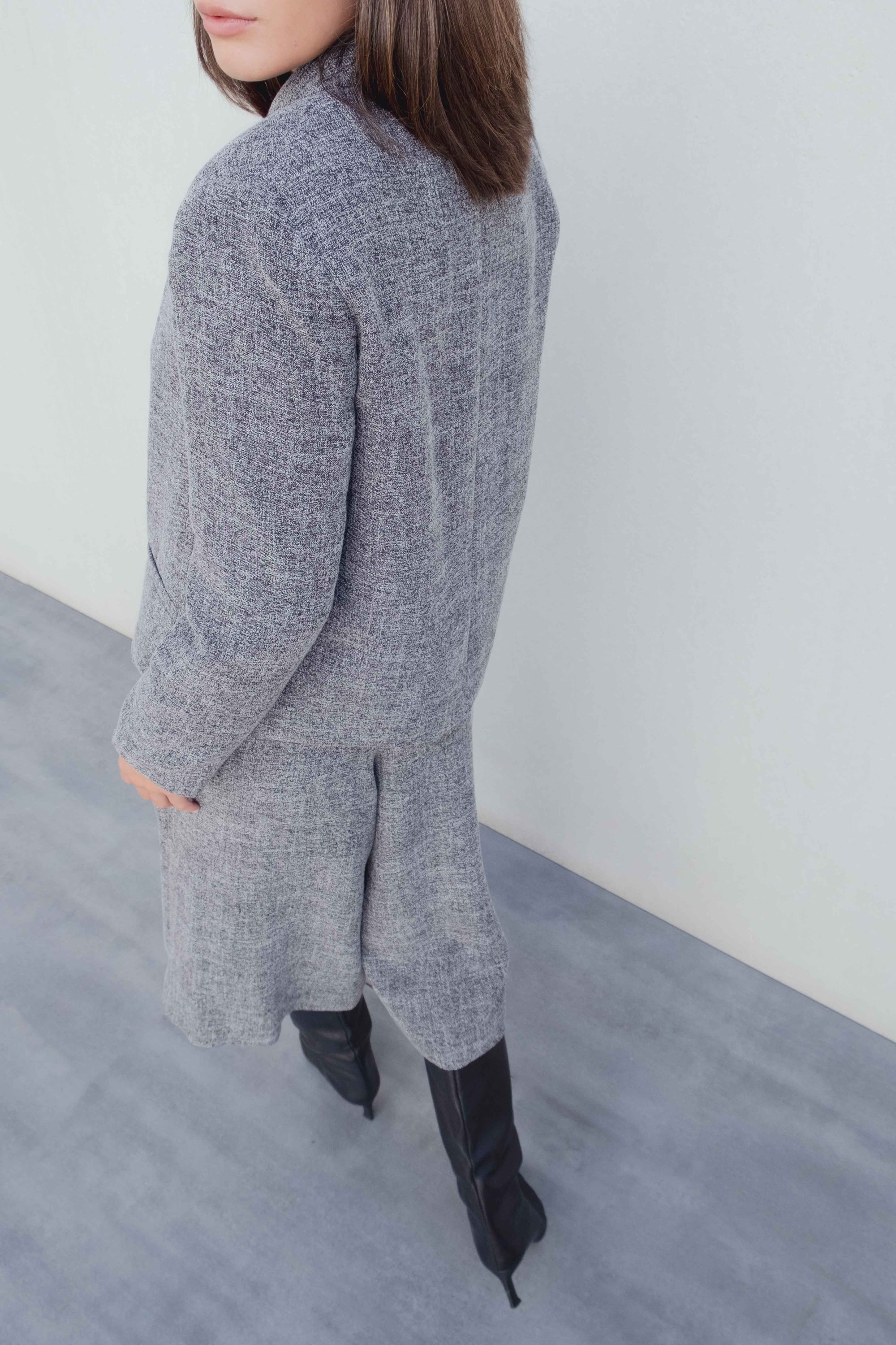 Vue arrière d'un modèle en blazer gris et Bermuda gris taille haute à pinces coupe large, avec des bottes noires stylées.