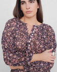 Femme avec un regard pensif, vêtue d'une blouse semi-transparente noire à fleurs, ajoutant une touche féminine à sa tenue. Blouse fleurie femme.