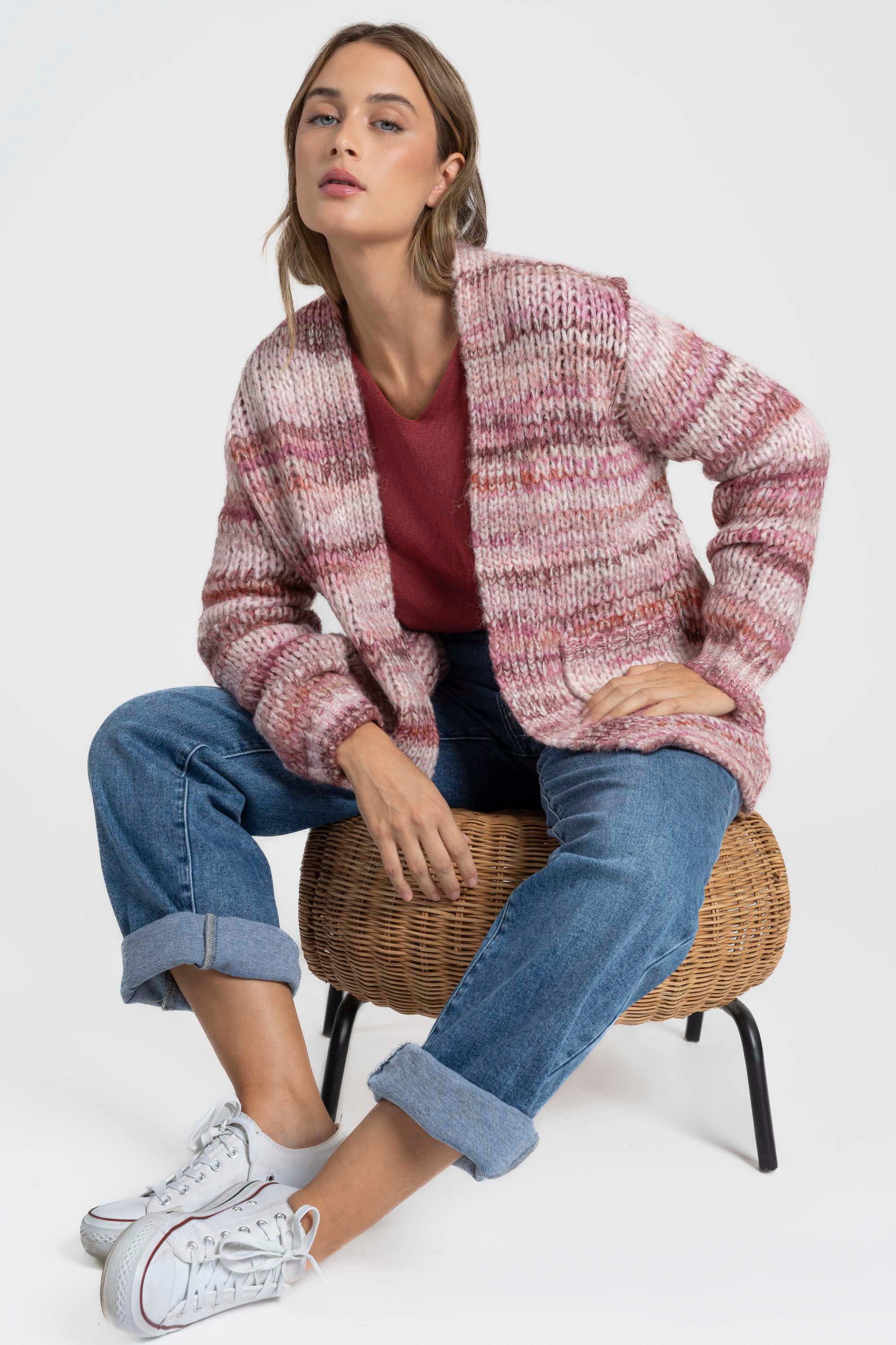Femme portant un cardigan court en maille, coloris rose et beige, assise sur une chaise en osier. Ce cardigan court femme en maille a une coupe moderne avec des poches.