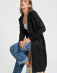 Modèle assise portant un cardigan long femme noir, en laine mélangée, avec des boutons et une structure fluide, parfait pour toute occasion.
