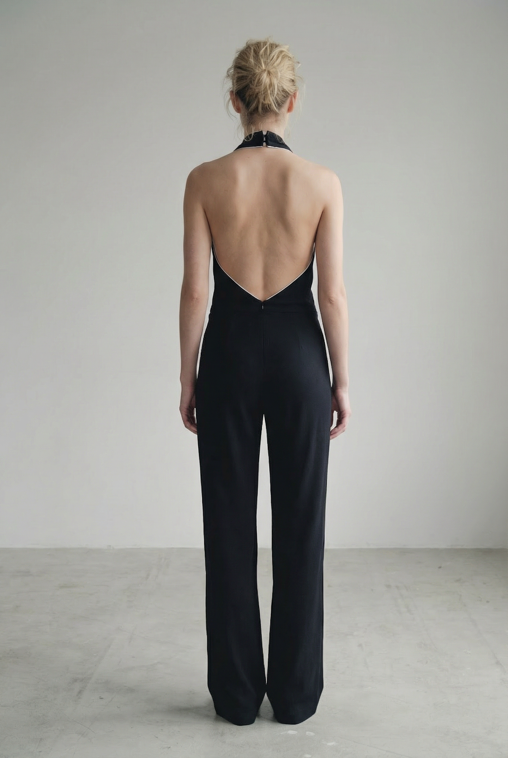 Vue arrière d'une femme portant une combinaison halter noire à nœud, dos nu et jambe large, illustrant l'allure et le style de la combinaison noire élégante femme.