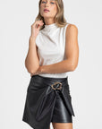 Femme élégante portant une jupe similicuir taille haute noire avec une boucle métallique, haut blanc.