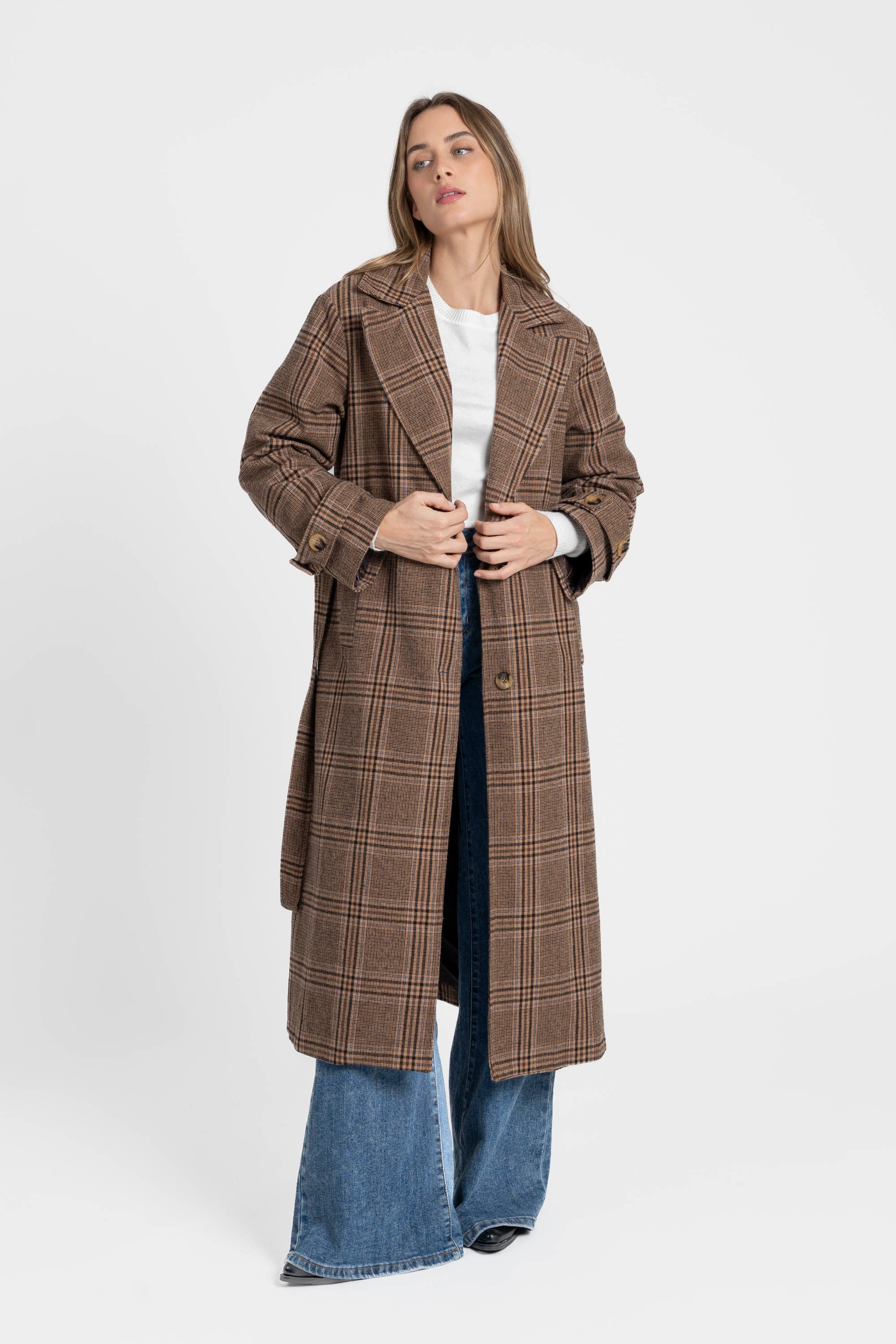 Manteau long femme en coton coupe ceinturée, présenté par une femme devant un fond blanc, en train de boutonner le manteau, style décontracté.