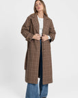 Manteau long femme en coton coupe ceinturée, présenté par une femme devant un fond blanc, en train de boutonner le manteau, style décontracté.